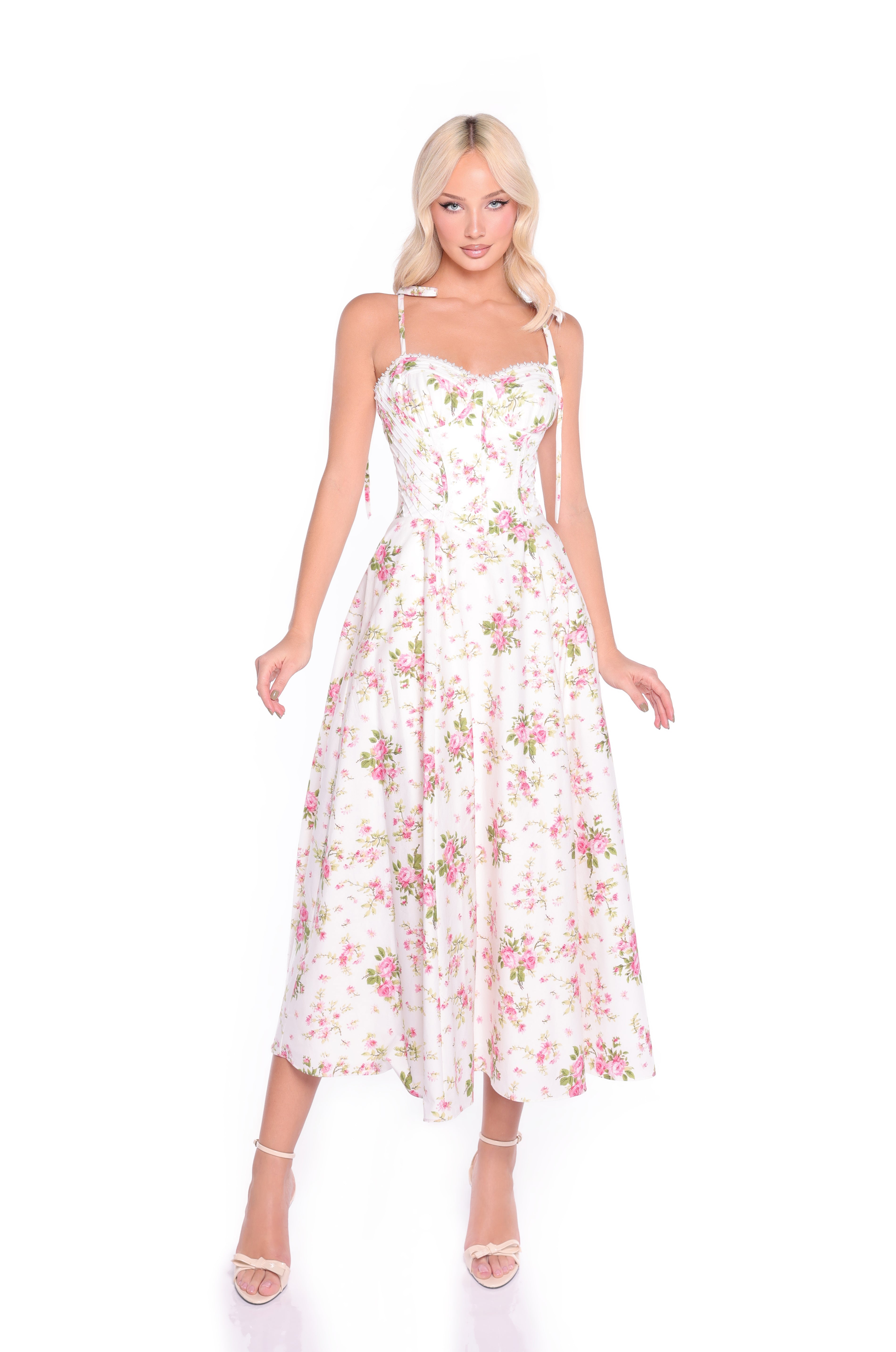 A woman wears the Ivory Blush Floral Midi Casual Dress: Cecilia, featuring a corset-style bodice and spaghetti straps, paired with strappy heels, standing against a plain white background.